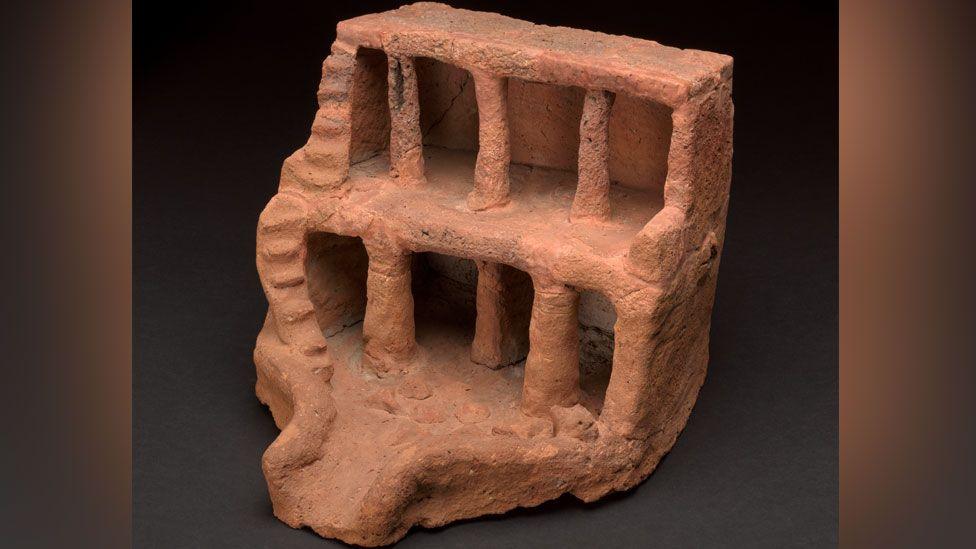 A view of a soul house made from lumpy light brown clay from the front. It is slightly off-centre to the viewer and shows a two storey open-fronted building, with pillars. Its ground floor has lumpen food offerings and an opening with a ramp for offerings to enter and exit. On its right side are steps rising up to the first and second floors. 