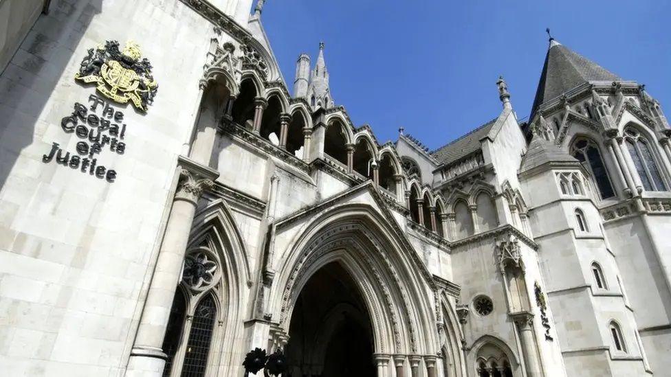 The outside of the Royal Courts of Justice in London. The picture is taken from a low angle, looking up at the imposing gothic facade of the building, which has a huge arched doorway and arched windows, similar to what you might see on a castle or a cathedral. 