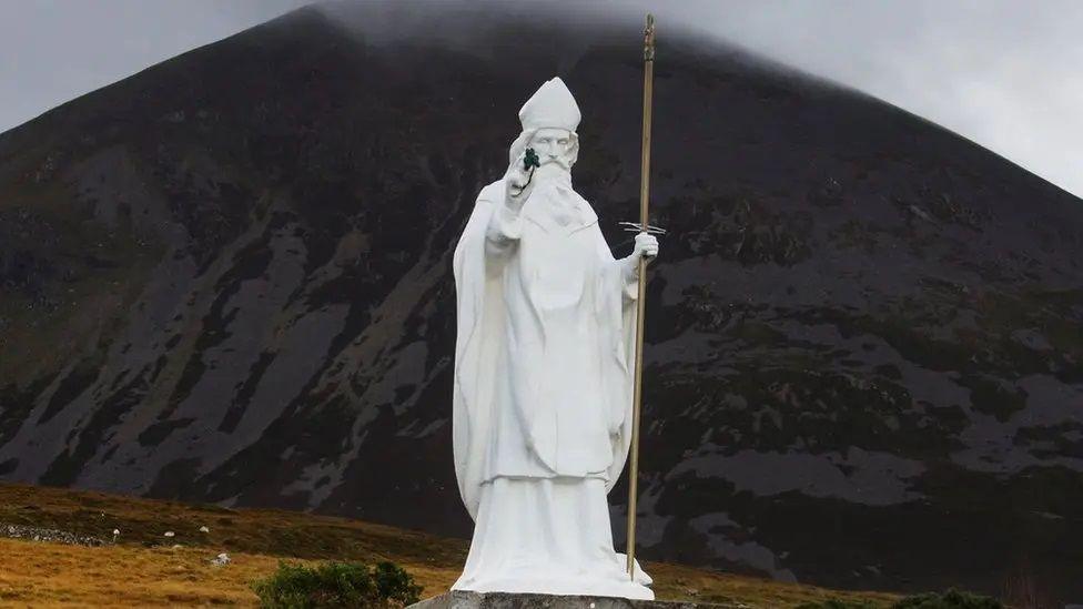 A white statue of St Patrick holding a shamrock standing in front of the Irish landscape in  Croagh Patrick in County Mayo, Ireland.