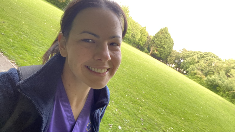 A selfie of a woman stood in a park. She has tied-back dark hair and is wearing purple nurse scrubs and a navy fleece. She is smiling and looking at the camera, with green grass and trees behind her.