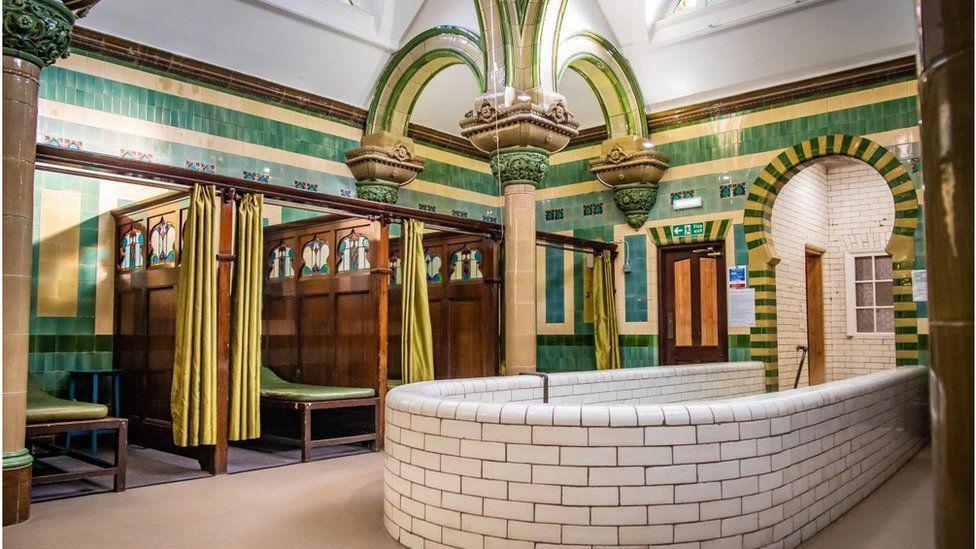 A highly decorated room with green and mustard tiles and ornate columns. There is a deep plunge pool in the foreground with white tiles. At the sides there are booths separated by wooden panels with decorations at the top and green privacy curtains.