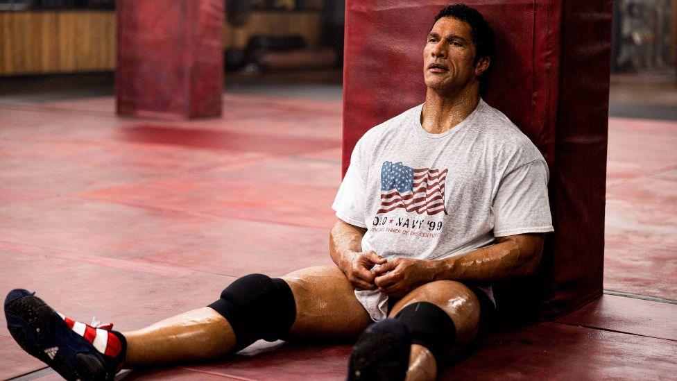 Dwayne Johnson as Mark Kerr in The Smashing Machine, he is sitting on the floor in a gym looking exhausted