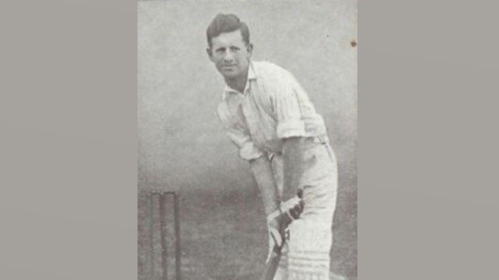 A black and white photo of Claude Ashton, in his cricket whites in a batting position, leaning over and holding a bat. A wicket can be seen on his right. He has short dark hair.
