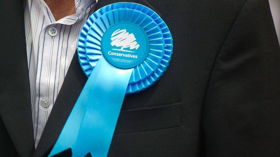 A man wearing a black suit over a white and blue striped shirt, with a Conservative rosette attached to him.