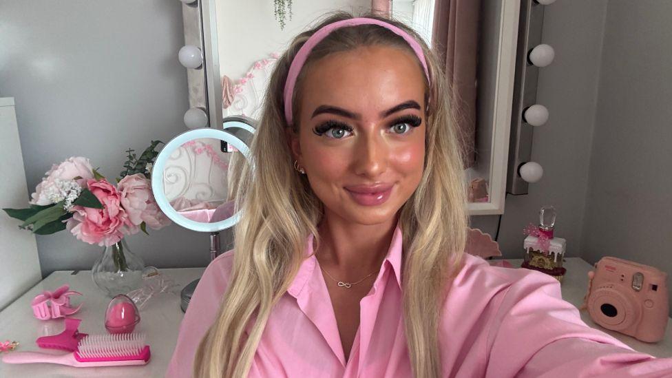 A young woman with long blonde hair wearing a pink shirt taking a selfie with beauty accessories in the background