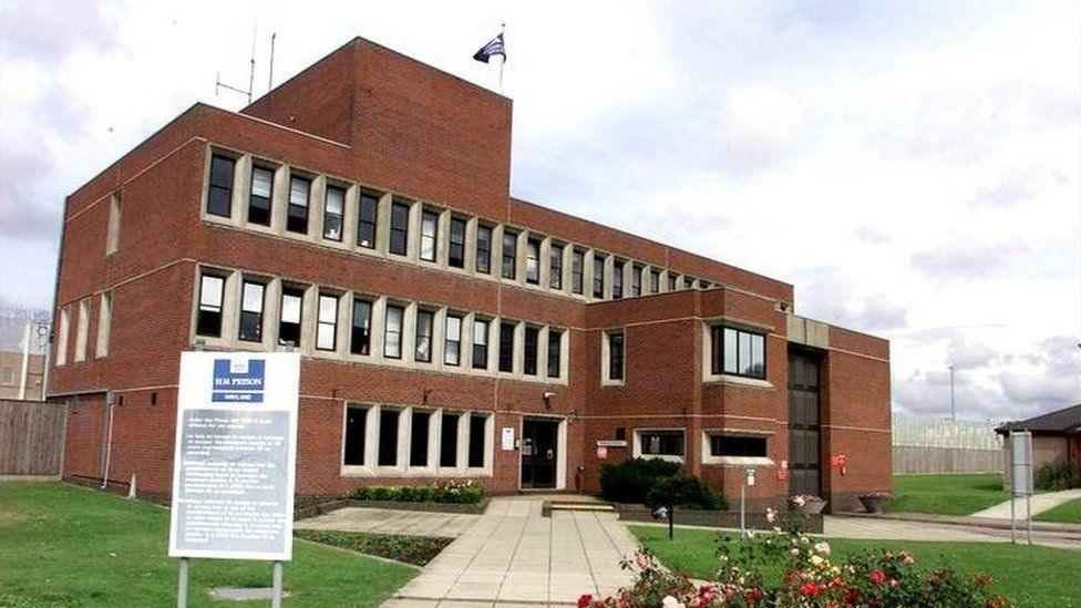 The entrance of HMP Wayland, which is a three-storey brick building with lots of windows. It is flanked by large fencing and has a grass lawn in front of it.