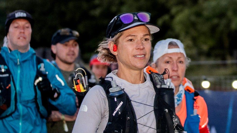 Stephanie Case is in the foreground of the photo. She wears a grey long-sleeved running top with a black hydration vest over it. She has a peaked black and grey cap on with sunglasses above the visor. One red earbud can be seen in her right ear. She is looking towards the right of the photo. A female runner is to her right and behind, and two male runners are behind her to the left. All three are slightly out of focus.