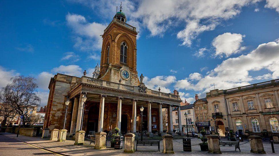 Front entrance of All Saints Church, with a pillared portico on the ground floor and a blue clock above. Also a figure of a man (Charles II) behind railings above the entrance.