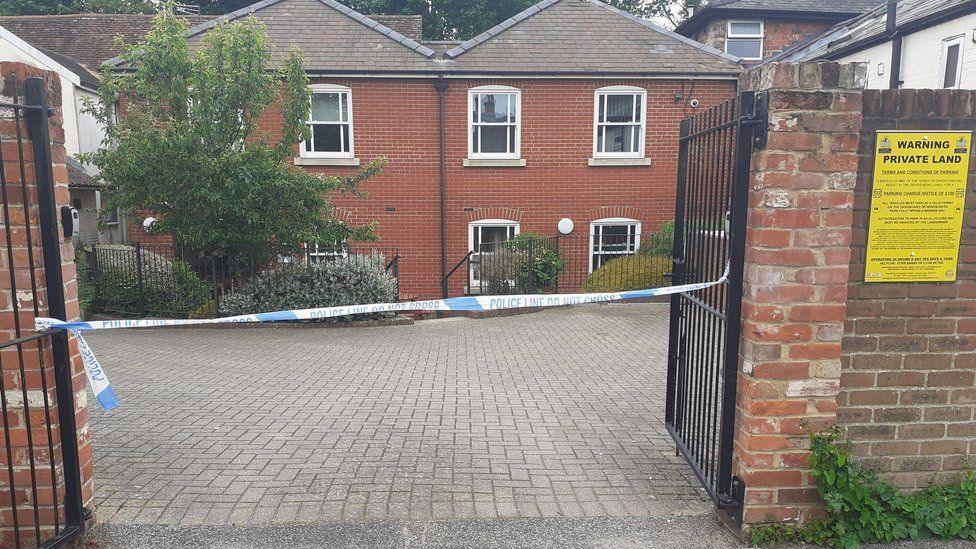 Police tape cordoning off the entrance to a courtyard outside some flats. The courtyard is made of grey bricks, while the flats themselves are two-storey and made of orange bricks.