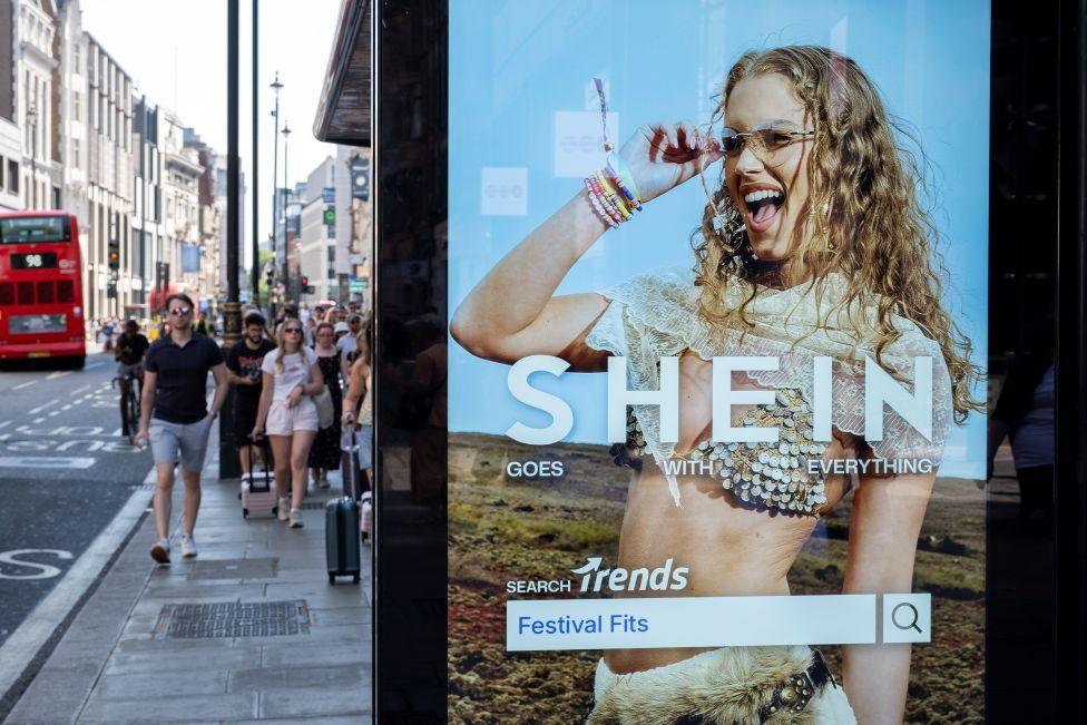 A Shein advert, showing a woman in a sequin bikini top and glasses, is displayed on a bus shelter as people walk down a busy high street