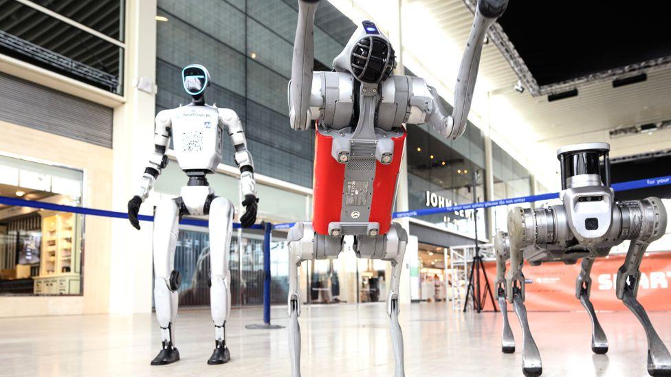 Three robots are in a cordoned area in a shopping centre. Two are almost humanoid and one is a little like a dog
