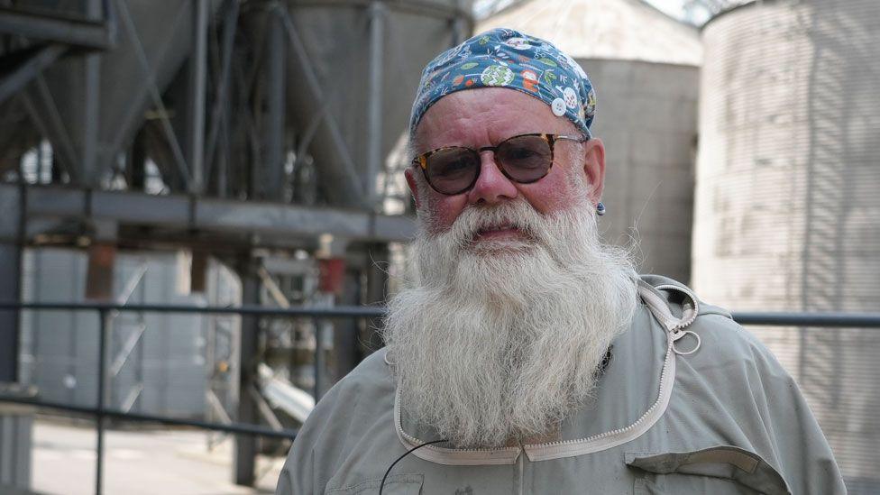 Beekeeper Steve Barrett wearing a grey beekeeping suit, but without its helmet. He has a long white beard and is wearing dark glasses and scarf on his head. Behind him are silver coloured buildings.