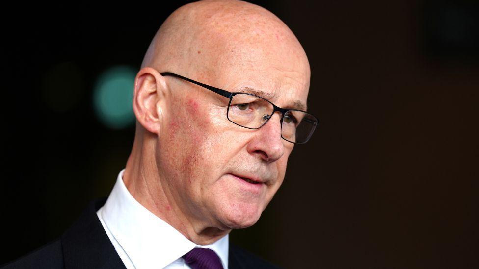 A close-up image of John Swinney, a bald man with glasses with a dark suit and white shirt