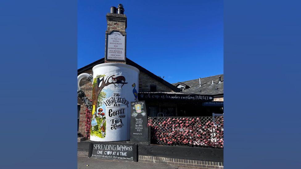 A large mug decoration on the chimney breast of a redbrick building. The mug is white with a floral handle and is painted with an image of Alice in Wonderland looking at a tree. There is a grinning cat in the tree and a bird to one side. The mug is advertising the name of the pub and coffee shop