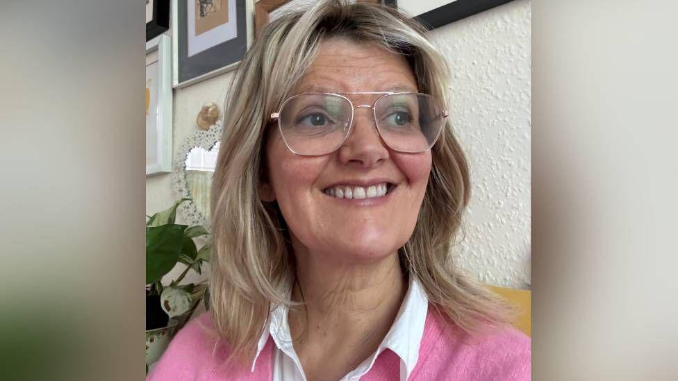 Iola Ynyr is smiling as she looks just away from the camera. She has long blonde hair and is wearing glasses, a white blouse and a pink jumper.