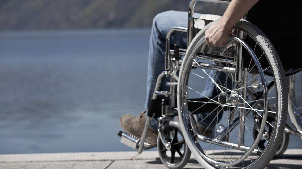 A stock close-up image of a person in a wheelchair showing a man's hand on a wheel rim and a foot in brown suede shoe on a footrest. The wheelchair is on a pavement overlooking a body of water.