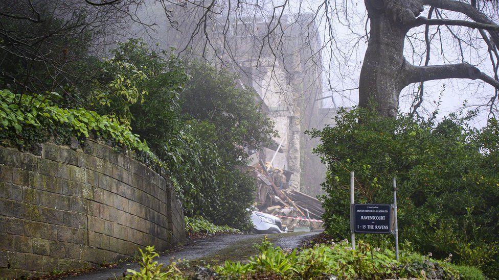 Steep driveway leading up to a building the front of white van is at the edge of red and white tape across the entrance with and a large pile of stone rubble and building debris.