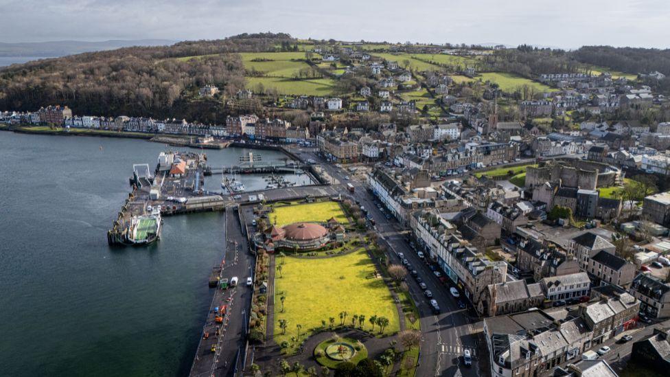 An aerial view of Rothesay. Rows of buildings divided by streets stretch out to the right, while a hill rises above the town on the far side of the picture. There is a harbour on the left side of the picture, next to a grassy area by the shoreline.  