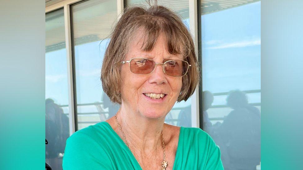 Christine Murton is in the middle of the image looking towards the camera. She has short brown hair and is wearing glasses and a green T-shirt.