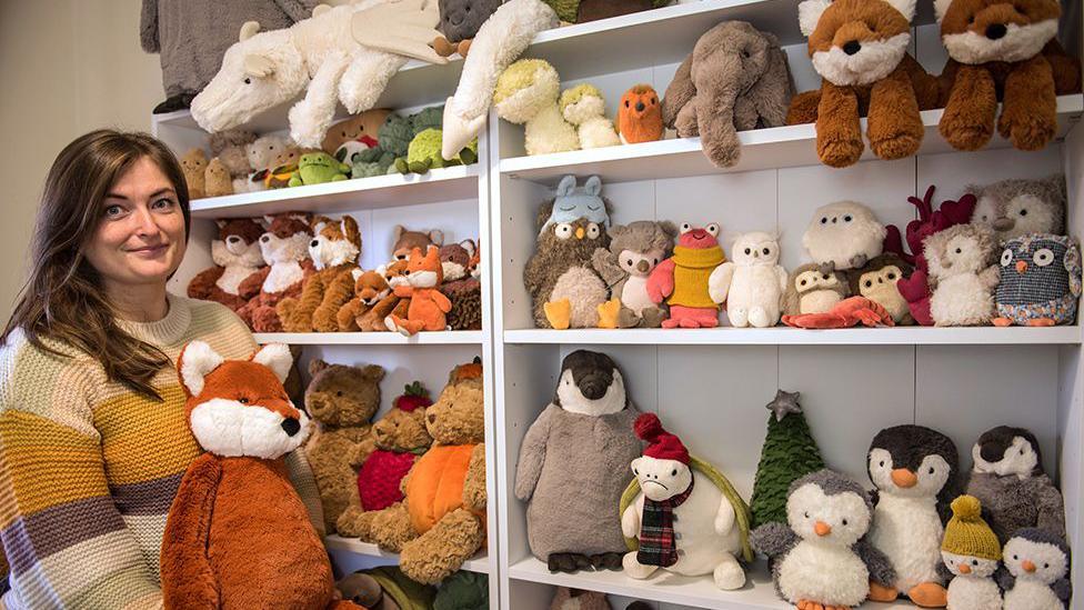 Amanda Hope stands holding a toy fox next to her collection of Jellycat toys stacked on shelves