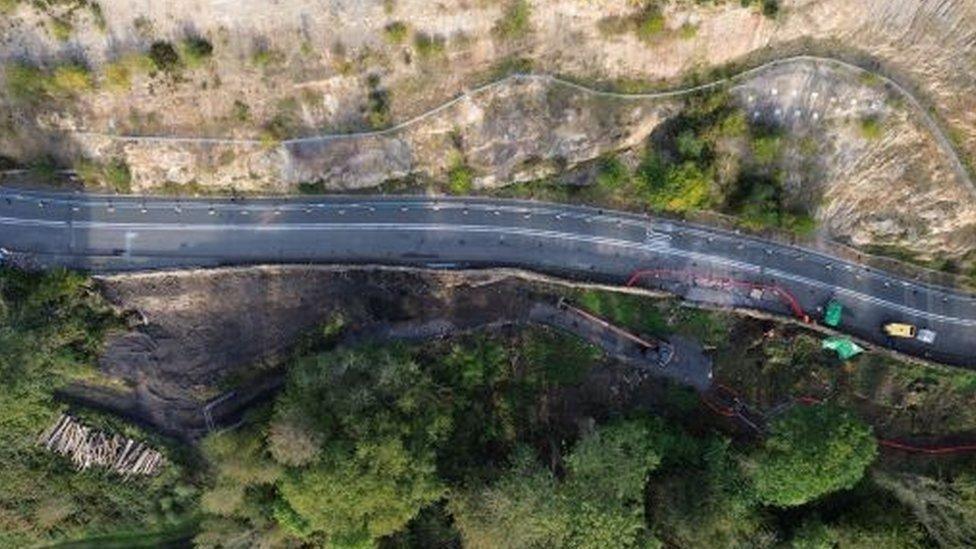 Aerial view of a road snaking into a gorge from left to right, with repair crews carrying out work to the lower wall and bank.