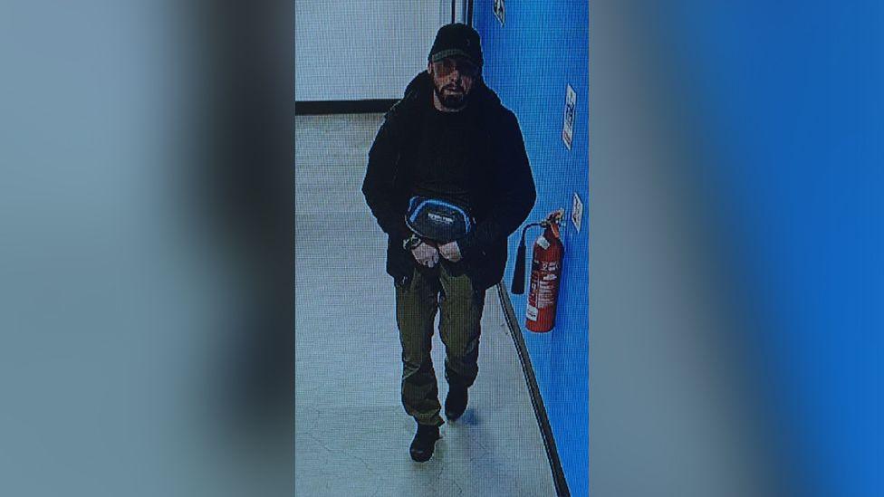 A CCTV image of James Clacher. He wears dark clothing and carries a bag while walking along a hallway