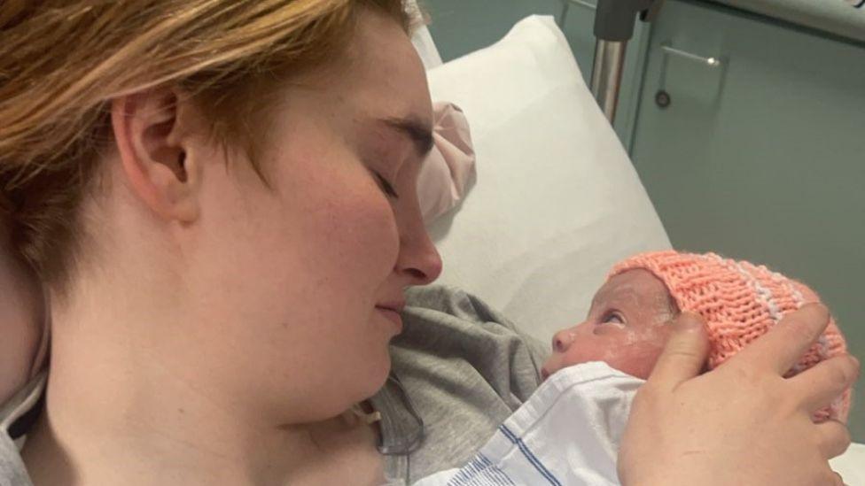 A mother, with light hair, holds a newborn baby wearing an orange knitted hat in a close-up shot taken in a hospital