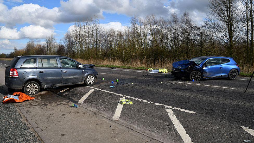 An otherwise empty main road with a grey car on the left and a blue car on the right, facing each other across the carriageway. Both cars have their fronts smashed in and there is debris across the road.