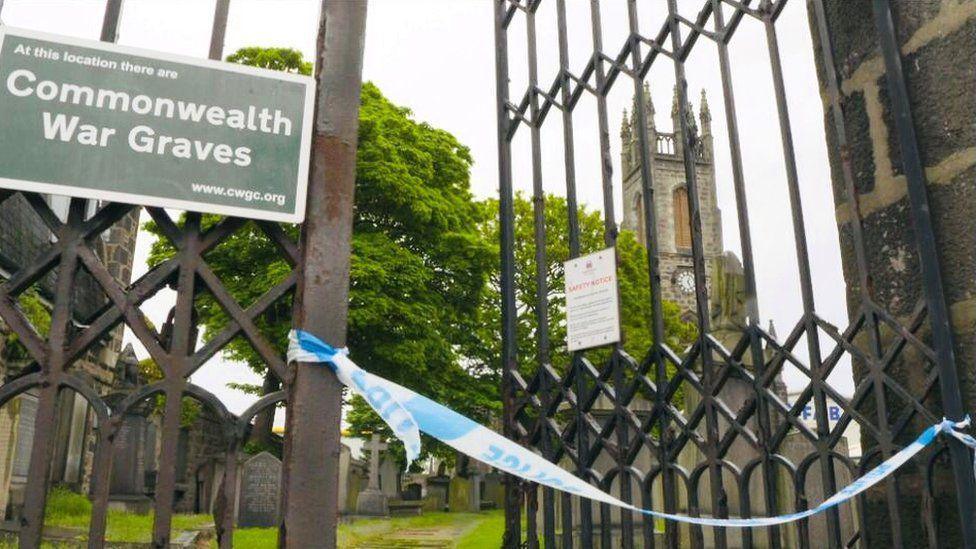 Iron gates outside church graveyard, with sign saying 'At this location are Commonwealth War Graves', one of the halves of the gates is ajar, with blue and white police tape across the opening, and graves and a church in the background.