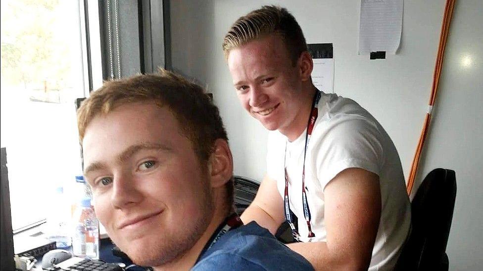 Guto and Robin Evans sit next to each other in a small office with a window. They are both turned to look at the camera and are smiling. They are wearing t-shirts and both have a lanyard around their necks. A keyboard and the edge of a screen can be seen to the left of the picture.