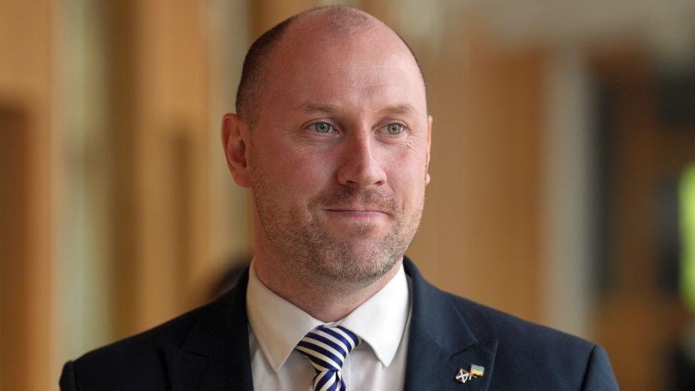 Neil Gray, a bald man with ginger stubble, looks off camera to his left. He is wearing a navy suit jacket, white shirt and striped tie