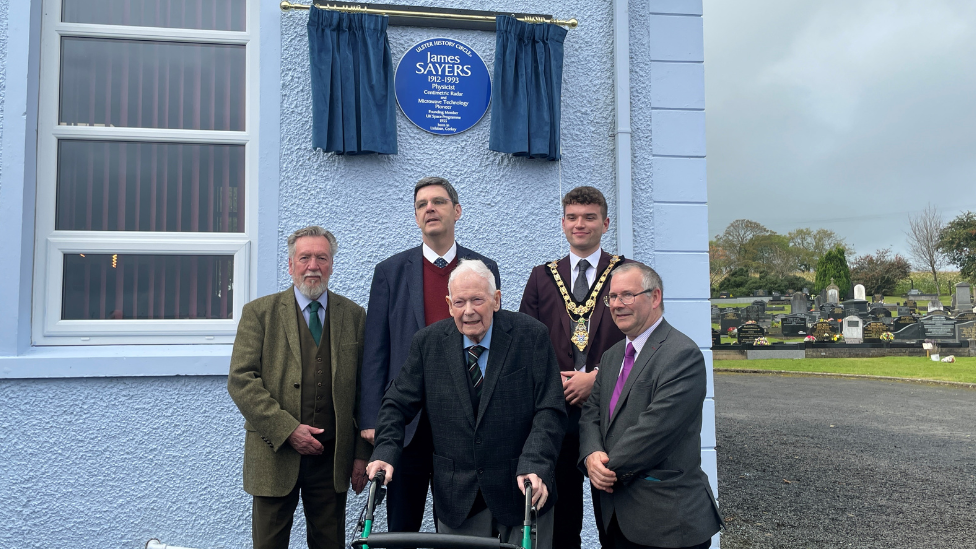 (L-R) Chris Spurr from Ulster History Circle, Rev Kenneth Henderson, Rev Robert Hanna, Mayor Ciarán McQuillan from Causeway Coast and Glens Borough Council and Dr David Hume from Ulster History Circle - all dressed in suit jackets, pictured under the plaque which is on side of a grey building, graveyard visible in the background