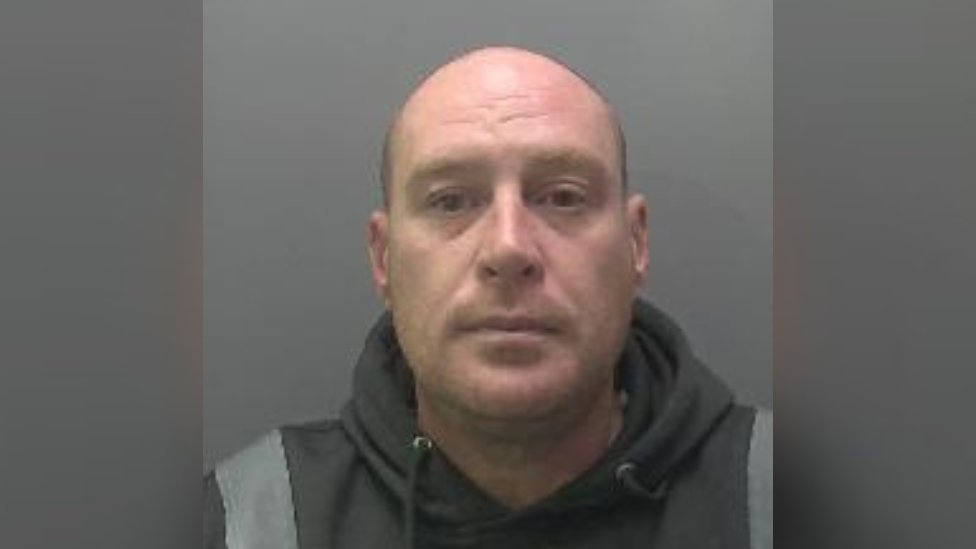 Custody photo of Darren Wyer, who is standing in front of a grey background. He has no hair and is looking directly at the camera. He wears a black hoodie with wide grey stripes on the shoulders. 