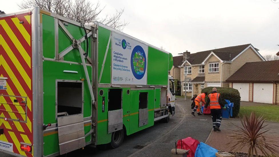 A green recycling collection truck is parked at the side of the road with holes in the side of it for the disposal of different materials. Council workers on the pavement collect the sacks from driveways with houses in the background.