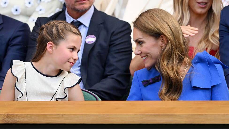 Princess Charlotte and the Princess of Wales sit front row in the royal box at Wimbledon. Princess Charlotte wears a white dress and smiles at her mother, who wears a blue dress, and is smiling back at her.