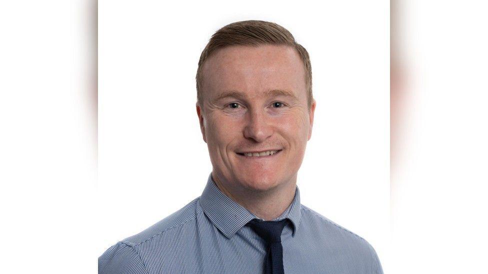 A head-and-shoulders shot of a man with short slicked-back ginger hair wearing a blue shirt and a navy tie. He is smiling into the camera and there is a white background