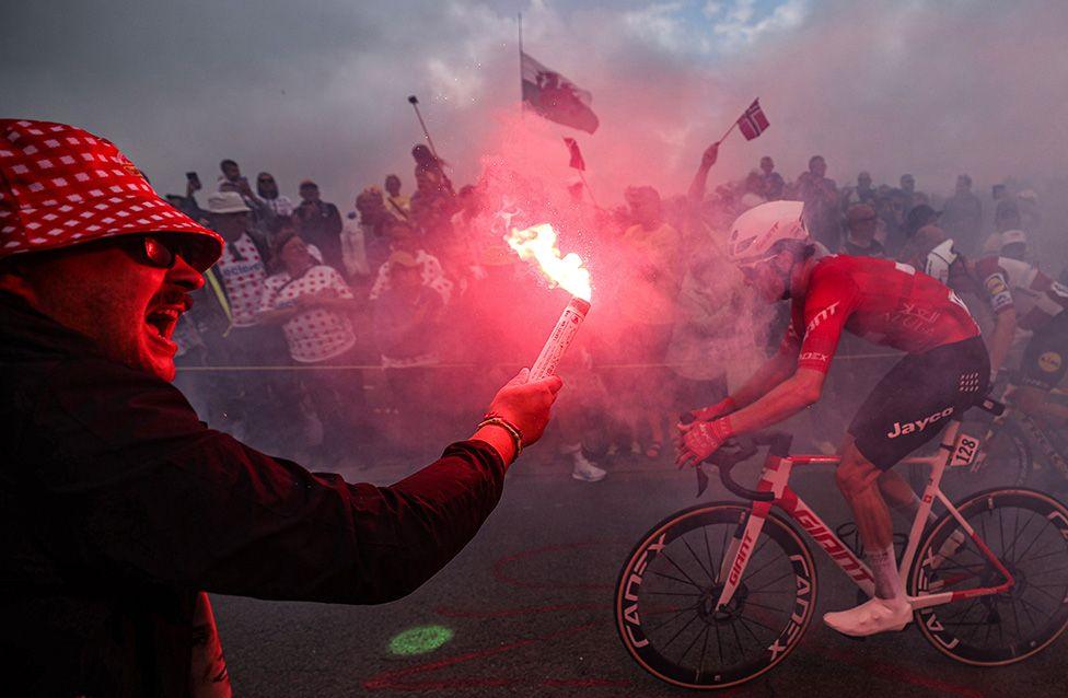 A spectator waves a flare as Jayco AlUla rider Mauro Schmid rides an ascent in the final kilometres of the second stage of the Tour de France.