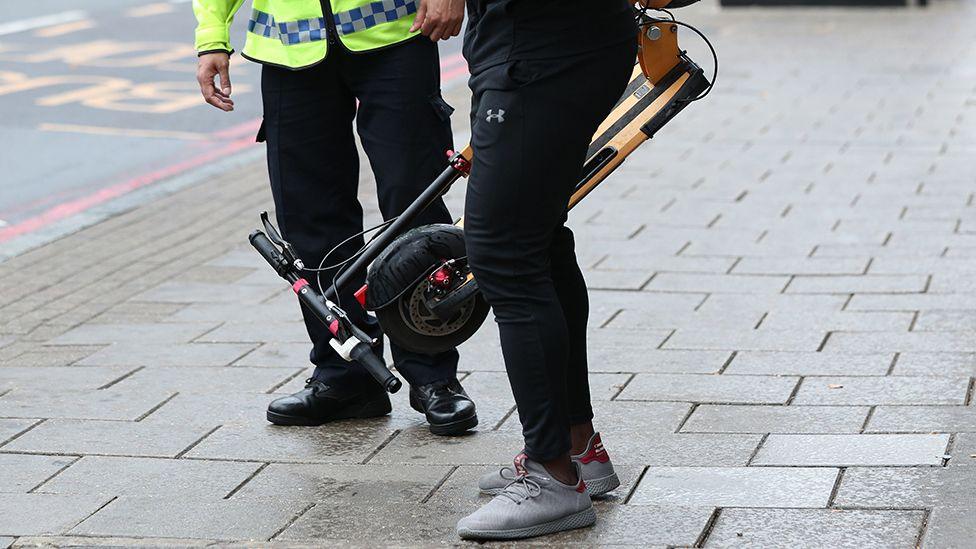 A man holding an e-scooter stood next to a police officer in a high-visability jacket. Only their legs are visible