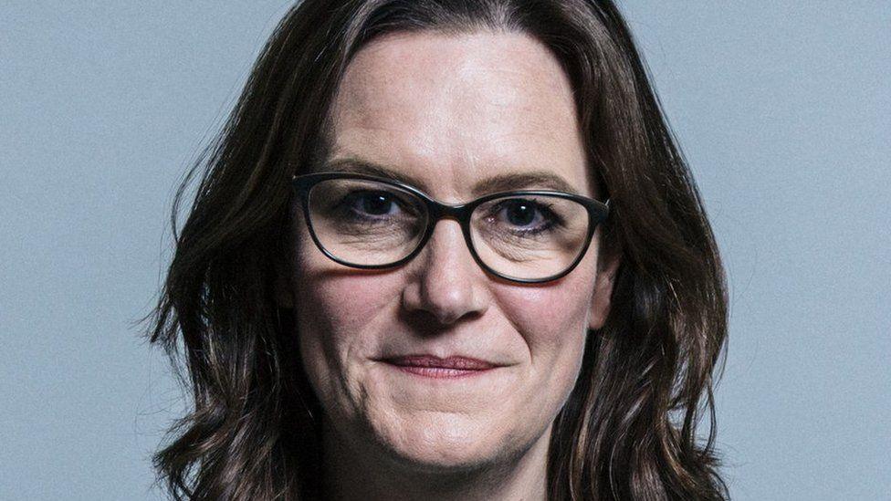 A close up of Rebecca Harris's face, who has brown hair and is wearing darkly-coloured framed glasses. She is standing inside, with a slightly-blue coloured background.