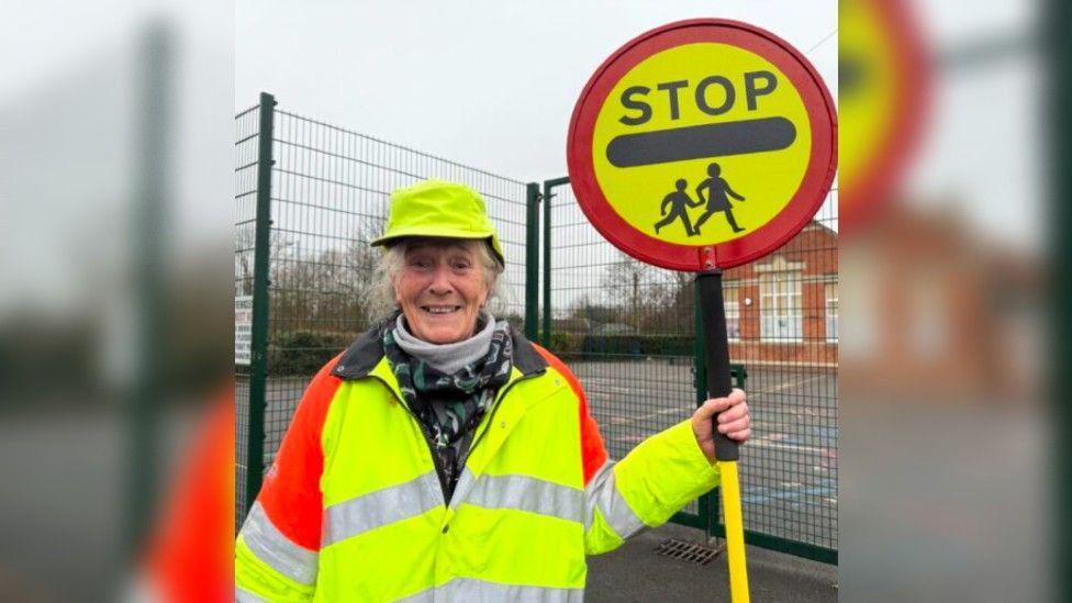 Heather standing outside on a dull cloudy day in her high vis jacket and holding her lollipop stick, in front of a school playground