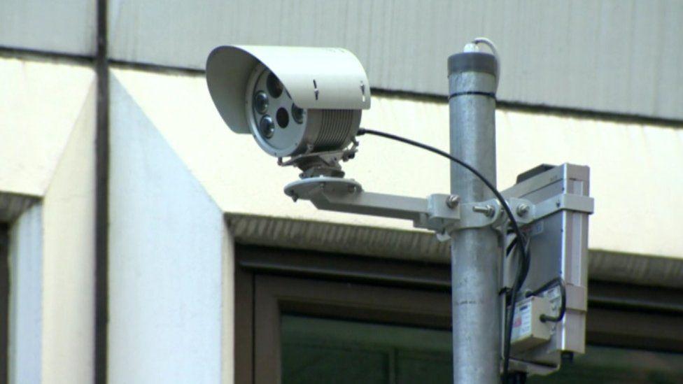 Grey multi-lens security camera mounted on a pole with a control box on the back. A window is visible behind.