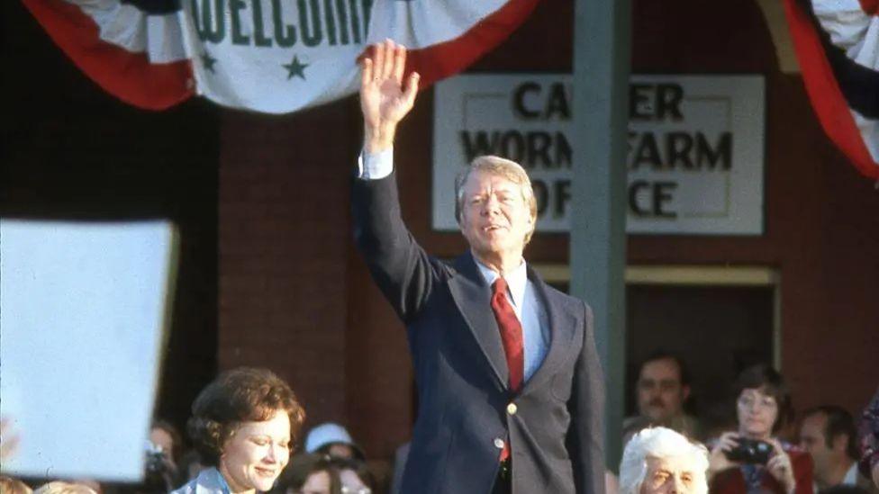 Carter holding a hand up and smiling as he stands above a crowd that is surrounding him