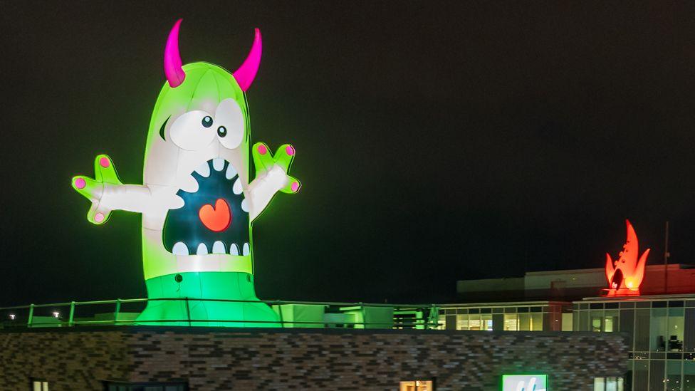 A green inflatable monster with a gaping mouth and pink horns stands on the top of a building while in the distance, another monster, with orange tentacles and a wide mouth, can be seen