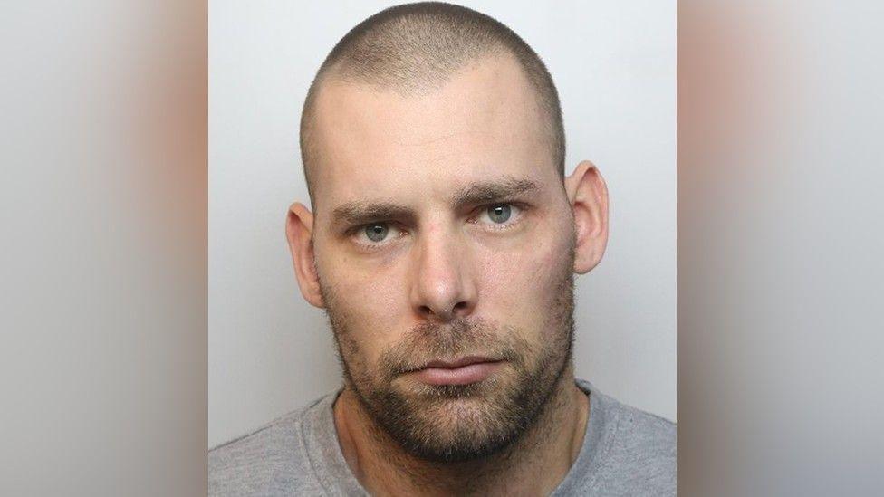 A mugshot of Damien Bendall. He has short, shaved hair and a lightly grown beard and moustache. He has green eyes and is wearing a grey T-shirt.