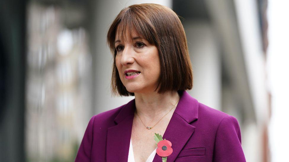 Rachel Reeves, with dark hair and wearing a purple suit, speaks with a blurred out concrete structure behind her 