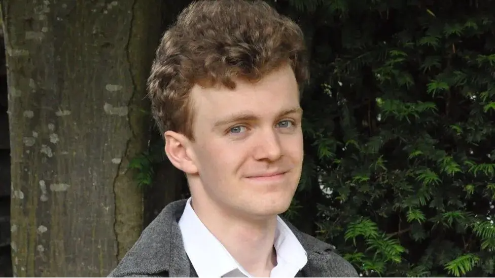 Sam Carling smiles and looks directly at the camera. He is photographed outside in front of green foliage and is wearing a white shirt and grey jacket. 