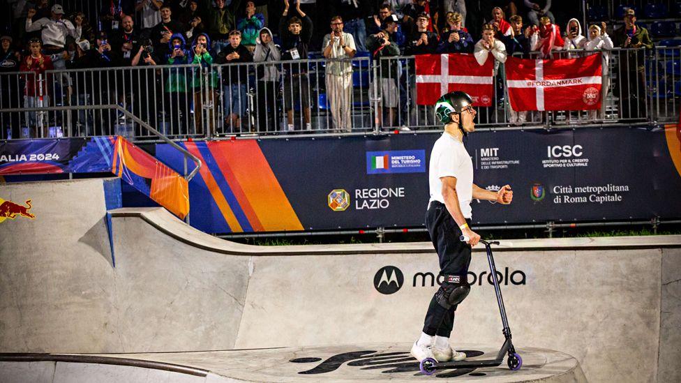 A side-profile shot of Jayden Sharman roaring at the skatepark, while people in the crowd cheer in the background.