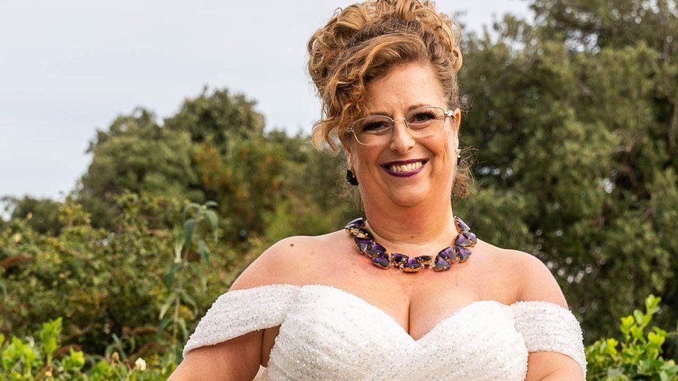 Sarah Wilkinson wearing glasses, an off-the shoulder white beaded dress, a purple stone necklace, her fair hair swept up into a bun with curls across her right forehead and smiling at the camera against trees in the background