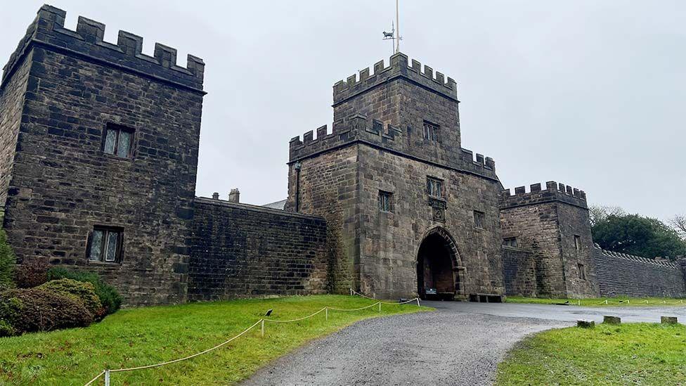 External image of Hoghton Tower's walled entrance with an arched gateway. The stone castle with grassed areas to the front has blackened stone