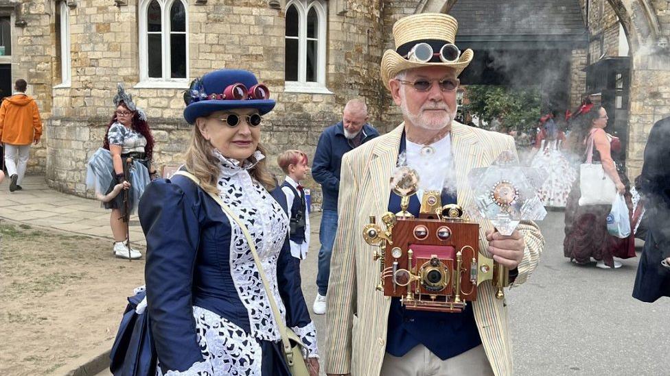 Two people attending the festival, with a man on the right wearing a cream and blue suit with an old Victorian camera and steam coming out of it. He wears a cream top hat and sunglasses. A woman on the left has a blue hat and sunglasses and is wearing a Victorian style blue and white long dress